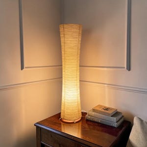 Pode incluir: Um candeeiro de pé alto e cilíndrico com um abajur de papel bege claro texturizado que emite um brilho quente e suave. O candeeiro apresenta um design esguio e ligeiramente cónico, posicionado verticalmente sobre uma superfície plana.