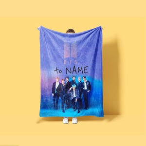 May include: A rectangular fleece blanket featuring a blue and purple gradient background. The top displays the BTS logo and the text "to NAME" in black. Below, a group photograph of seven individuals in dark clothing is printed on the fabric.