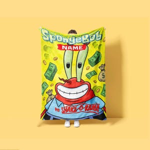 Op de afbeelding: Een gele fleece deken met een afbeelding van Meneer Krabs uit SpongeBob SquarePants. Het ontwerp bevat de tekst SpongeBob, NAME en in SHAKE-O-RAMA, omringd door illustraties van groene bankbiljetten, gouden munten en geldzakken.