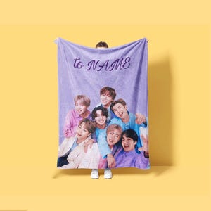 May include: A rectangular purple fleece blanket featuring a printed group photograph of seven individuals. The text "to NAME" is printed in a dark purple cursive font at the top center of the blanket. The fabric has a soft, textured appearance.