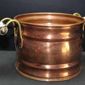 May include: A round copper bucket with a polished metallic finish and two curved brass handles. Each handle features a white ceramic grip with blue floral patterns. The bucket has a wide opening and a slightly flared base with horizontal ridges.