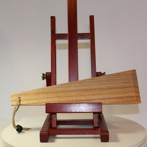 May include: A light brown wooden wedge with a visible grain pattern, featuring a tan cord attached to the narrow end with a small black bead. The wedge is positioned horizontally on a dark reddish-brown wooden easel stand.