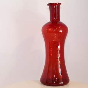 May include: A tall, translucent red glass vase with a slender neck and a wide, flared base. The vessel features a smooth, polished surface that catches the light, creating subtle highlights along its curved, elongated silhouette and rounded body.