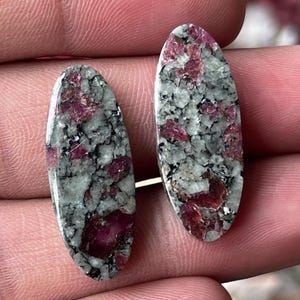 Op de afbeelding: Twee ovaalvormige cabochons met een gespikkeld patroon van lichtgrijze en witte mineraalmatrix met ingebedde diepmagenta en donkerrode kristallijne insluitingen. De stenen hebben een glad, gepolijst oppervlak en een langwerpige ovale vorm.