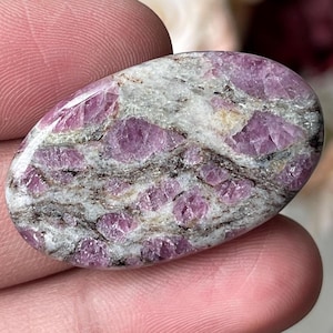 May include: An oval-shaped polished gemstone featuring a mix of vibrant pink crystalline inclusions embedded within a light gray and white matrix, accented by thin, dark, irregular mineral veins running across the smooth, glossy surface.