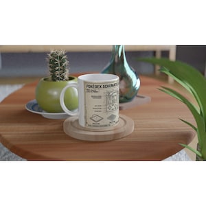 Pode incluir: Uma caneca de cerâmica branca com uma estampa gráfica em tom bege de um esquema de Pokédex. O design inclui desenhos técnicos de linhas de um dispositivo portátil, peças rotuladas e o texto Pokédex Schematic e Total Pokémon Registered 151.