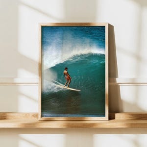 Op de afbeelding: Een ingelijste kunstdruk met een surfer op een grote, krullende blauwe oceaangolf. De surfer staat op een longboard en draagt een oranje zwembroek, terwijl de top van de golf breekt met wit zeeschuim op de achtergrond.