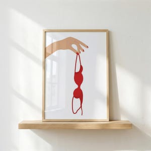 Könnte beinhalten: Ein gerahmter Kunstdruck mit einer minimalistischen Illustration einer Hand mit roten Fingernägeln, die einen leuchtend roten BH an einem der Träger hält. Das Kunstwerk ist vor einem schlichten weißen Hintergrund in einem hellen Holzrahmen.