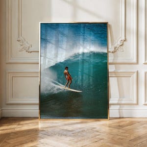 Könnte beinhalten: Ein gerahmter vertikaler Wandkunstdruck, der einen Surfer auf einer großen, tiefblauen Meereswelle zeigt. Der Surfer steht auf einem hellen Surfbrett und trägt eine orangefarbene Badehose, während sich der Wellenkamm in einer kraftvollen Bewegung krümmt.