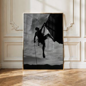Könnte beinhalten: Ein schwarz-weiß gerahmter Wandkunstdruck, der die Silhouette eines Felskletterers an einer steilen Felswand zeigt. Der Kletterer hängt an einem Seil vor einem bewölkten Himmel, mit dunklen Berggipfeln entlang der unteren Horizontlinie.