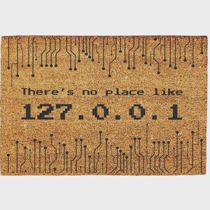 Puede incluir: Un felpudo rectangular de fibra de coco color canela con un patrón de circuito impreso negro en los bordes superior e inferior. El centro de la alfombra muestra el texto negro "There's no place like 127.0.0.1" en una fuente estilo informático.