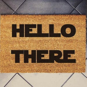 Pode incluir: Um capacho retangular de fibra de coco em tom castanho natural com o texto HELLO THERE impresso em letras maiúsculas pretas e em negrito no centro. O tapete tem uma textura fibrosa e um design simples e limpo para a entrada.