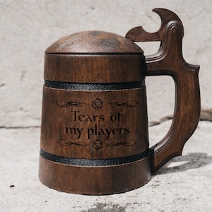 Puede incluir: Una jarra de cerveza de madera de color marrón oscuro con tapa y asa. La jarra presenta dos bandas horizontales negras y el texto grabado "Tears of my players" centrado en el frente, flanqueado por pequeños iconos geométricos.