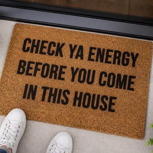 May include: A rectangular brown coir doormat with black text that reads, "CHECK YA ENERGY BEFORE YOU COME IN THIS HOUSE." The mat has a coarse, natural fiber texture and is positioned on a light-colored floor surface near a doorway entrance.