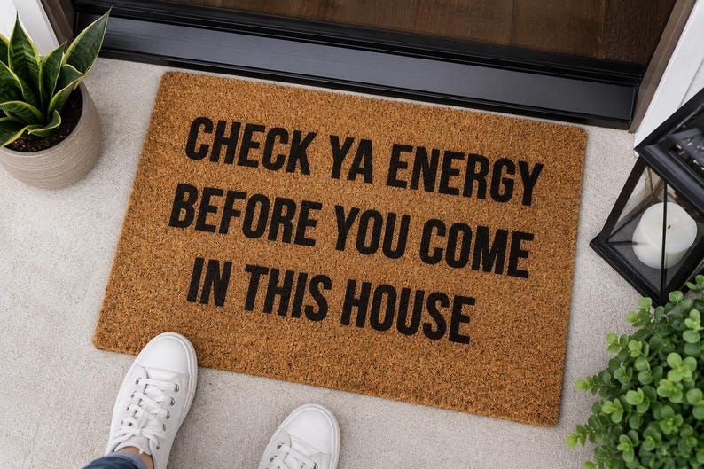 May include: A rectangular brown coir doormat with black text that reads, "CHECK YA ENERGY BEFORE YOU COME IN THIS HOUSE." The mat has a coarse, natural fiber texture and is positioned on a light-colored floor surface near a doorway entrance.