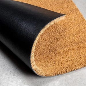 May include: A rectangular doormat featuring a coarse, tan-colored natural fiber top surface and a smooth, solid black rubberized backing. The mat is partially folded over to reveal the contrast between the textured brown front and the black underside.