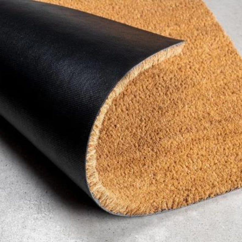 May include: A rectangular doormat featuring a coarse, tan-colored natural fiber top surface and a smooth, solid black rubberized backing. The mat is partially folded over to reveal the contrast between the textured brown front and the black underside.