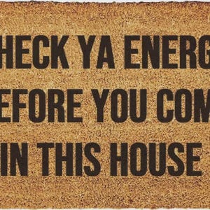 May include: A rectangular tan coir doormat with a rough, fibrous texture. The mat features bold, black, sans-serif text printed in the center that reads: CHECK YA ENERGY BEFORE YOU COME IN THIS HOUSE. The edges of the mat are straight and defined.