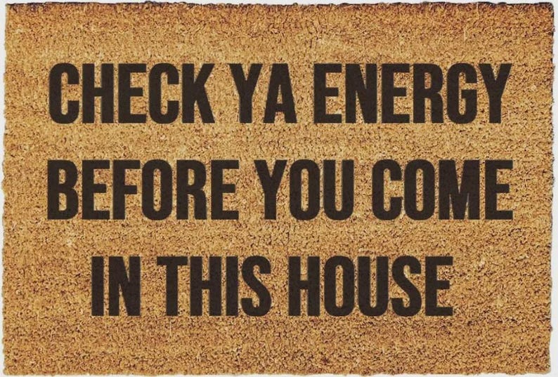 May include: A rectangular tan coir doormat with a rough, fibrous texture. The mat features bold, black, sans-serif text printed in the center that reads: CHECK YA ENERGY BEFORE YOU COME IN THIS HOUSE. The edges of the mat are straight and defined.