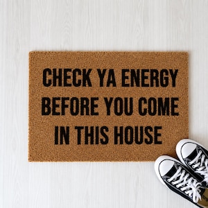 May include: A rectangular brown coir doormat with black text that reads: CHECK YA ENERGY BEFORE YOU COME IN THIS HOUSE. The mat has a coarse, fibrous texture and is positioned next to a pair of black and white canvas sneakers with white laces.