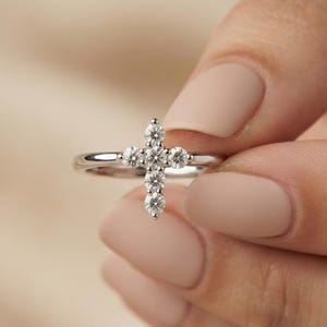 May include: A silver-toned ring featuring a cross-shaped centerpiece adorned with five small, clear, round-cut stones. The band is smooth and polished, with the cross design positioned vertically across the top of the ring's setting.