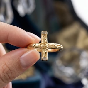 Könnte beinhalten: Ein goldener Ring mit einem Kruzifix-Design und einer zentralen Jesusfigur. Das Kreuz ist mit zwei kleinen, rechteckigen Diamanten im Baguetteschliff verziert, die an den oberen und unteren Enden des vertikalen Balkens eingefasst sind.