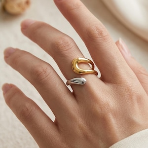 May include: A two-tone open ring featuring a polished gold-colored curved top section and a smooth silver-colored bottom section. The ring has a bypass design with rounded ends that do not meet, creating a continuous fluid shape on the finger.