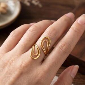 May include: A gold-toned open ring featuring two distinct teardrop-shaped wire motifs. The design consists of a smaller teardrop element on one side and a larger, layered teardrop element on the other, creating a continuous, fluid, and minimalist aesthetic.
