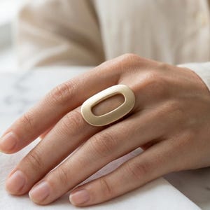 May include: A gold-toned ring featuring a large, horizontal, oval-shaped band with a smooth, matte finish. The ring has a central open cutout design, creating a bold, geometric look that sits prominently across the finger.