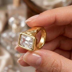 Peut inclure: Une bague dorée ornée d'une grande pierre précieuse rectangulaire transparente à facettes, montée sur un anneau en métal épais et poli. La monture présente une forme organique lisse qui encadre la pierre centrale, offrant un aspect audacieux.
