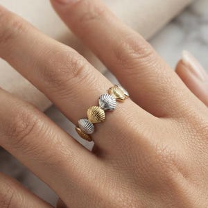 May include: A ring featuring a band composed of alternating silver-toned and gold-toned seashell motifs. The small, textured shells are arranged in a continuous row around the circumference of the band, creating a repeating pattern of metallic colors.