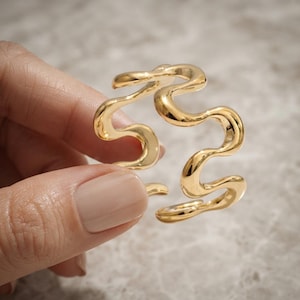 Op de afbeelding: Een goudkleurige open armband met een doorlopend, vloeiend, golvend of slangachtig ontwerp. Het metaal heeft een gepolijst, reflecterend oppervlak dat het licht opvangt en vloeiende rondingen creëert over de gehele structuur van het sieraad.