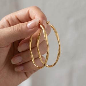 May include: A pair of gold hoop earrings featuring a distinct twisted, wavy texture. The earrings are circular in shape with a polished metallic finish, showcasing a continuous undulating design that creates a dynamic visual effect along the entire band.