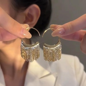 Puede incluir: Un par de pendientes de aro dorados con una sección superior semicircular incrustada con cristales claros y brillantes. De la base de cada semicírculo cuelga un fleco denso de cadenas doradas brillantes que cuelgan hacia abajo.