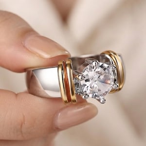 May include: A silver-toned ring featuring a large, round, clear faceted stone set in a six-prong setting. The band is wide and polished, accented on both sides of the central stone by two parallel, raised gold-toned bands that wrap around the ring.