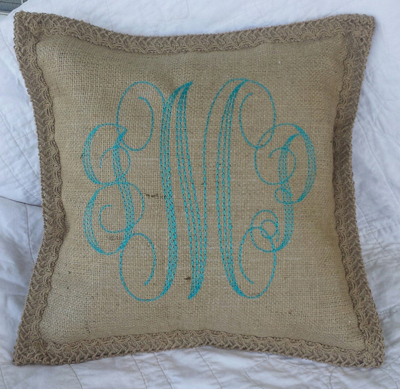 May include: A burlap pillow with a turquoise embroidered monogram. The pillow has a burlap trim.