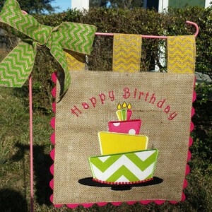 May include: A burlap birthday flag with a pink scalloped edge. The flag features a green and yellow cake with pink frosting and candles. The text "Happy Birthday" is embroidered in pink on the flag.