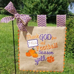 May include: A burlap garden flag with the text "Thank GOD for Fall and Football Season y'all". The flag features embroidered pumpkins, a paw print, and a purple and gold chevron bow. The edges are trimmed with orange.