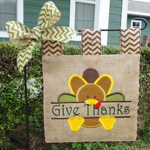 May include: A burlap Thanksgiving garden flag with a brown and green turkey design. The turkey is peeking over a banner that says "Give Thanks". The flag is attached to a black metal pole with a green and brown chevron bow.