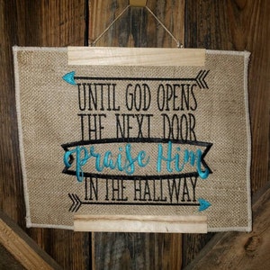 May include: A burlap wall hanging with a wooden frame. The hanging features black and white embroidery with the text "Until God opens the next door praise Him in the hallway". The text "praise Him" is embroidered in teal.