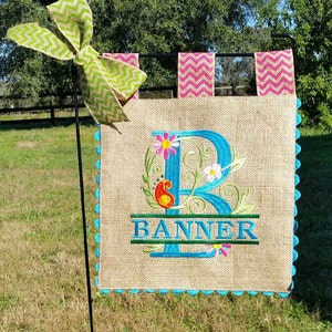 May include: A burlap garden flag with a blue letter "B" surrounded by colorful flowers and the word "BANNER" embroidered on it. The flag has a scalloped edge and is attached to a black metal stake.