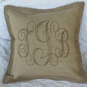 May include: A brown burlap pillow with a decorative monogram embroidered in brown thread. The pillow has a brown burlap trim.