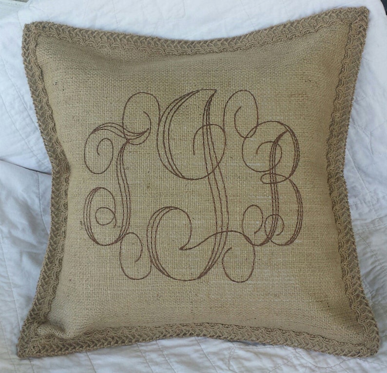 May include: A brown burlap pillow with a decorative monogram embroidered in brown thread. The pillow has a brown burlap trim.