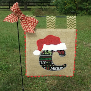 May include: A burlap Christmas garden flag with a red and white Santa hat and the letter C in black with the words "Jolly", "Christmas", "Love", and "Merry" around it.