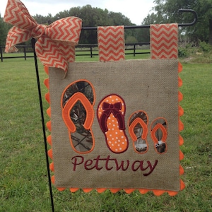May include: A burlap garden flag with a scalloped orange border. The flag features three embroidered flip-flops in camouflage, polka dot, and solid orange patterns. The name "Pettaway" is embroidered in maroon below the flip-flops.