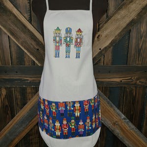 May include: Off-white apron featuring a colorful embroidered design of three nutcracker soldiers. The bottom of the apron has a blue band with a repeating pattern of nutcrackers in various uniforms. The apron has white straps.