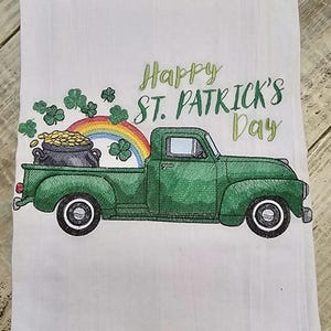 May include: White kitchen towel with an embroidered design featuring a green truck carrying a pot of gold, a rainbow, and shamrocks. The text "Happy St. Patrick's Day" is embroidered in green. The towel is likely made of cotton.