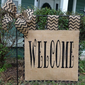 May include: A burlap welcome sign with black lettering and a black and white chevron patterned ribbon bow. The sign is attached to a black metal stand.