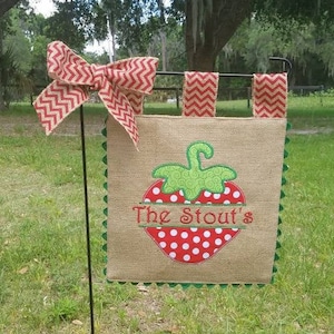 May include: A burlap garden flag featuring a red and white zig-zag bow and matching accents. The flag displays a red strawberry with white polka dots and the text "The Stout's" in green and red.