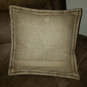 May include: A square burlap pillow with a black trim. The pillow is sitting on a brown leather couch.